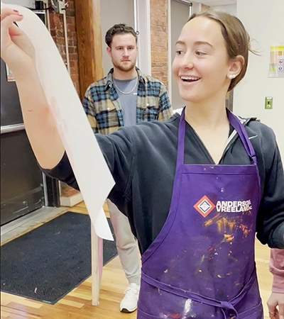 Young woman in a colorful apron smiling while holding up a painted sheet of paper, with a man in the background observing.