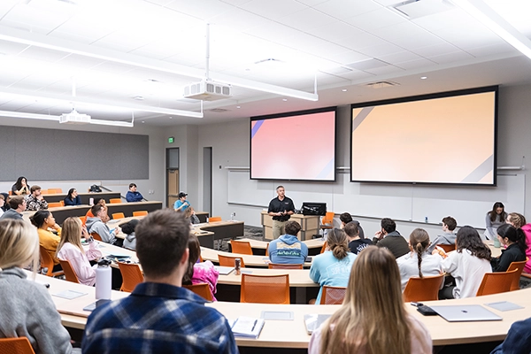 A modern classroom setting with a lecturer presenting to an engaged audience, featuring large screens and a circular seating arrangement.