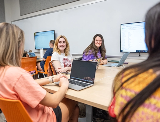 Four students collaborating in a classroom setting, working on laptops and engaged in discussion.