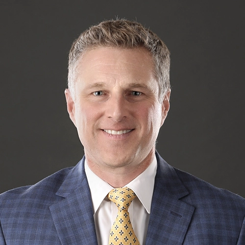 Professional headshot of a man in a blue suit and yellow patterned tie against a dark background.
