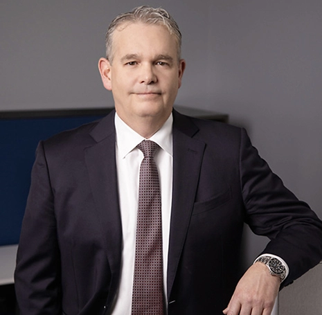 A professional headshot of a middle-aged man in a suit, standing confidently with his hand on a desk.