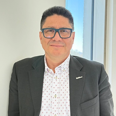 Smiling man wearing glasses and a dark blazer over a patterned shirt, standing indoors near a window.