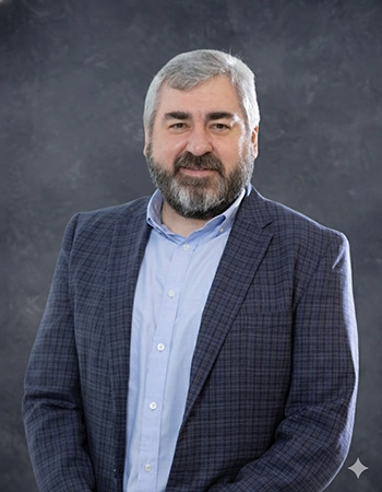 Portrait of a man with a beard, wearing a checkered blazer and light blue shirt, against a gray backdrop.