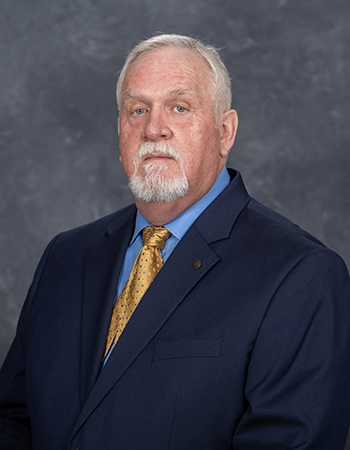 Professional portrait of a man in a suit with a light blue shirt and yellow tie. He has a white beard and short hair, posing against a gray background.