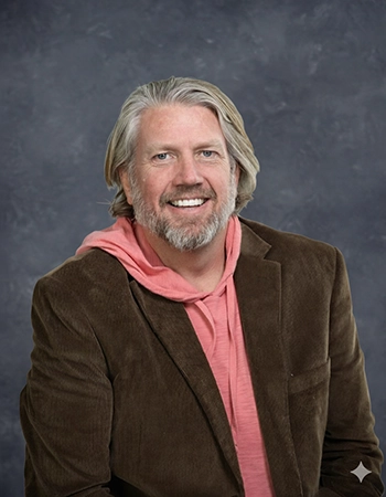 A friendly middle-aged man with long gray hair and a beard, wearing a brown blazer over a pink hoodie, smiles confidently against a textured gray backdrop.