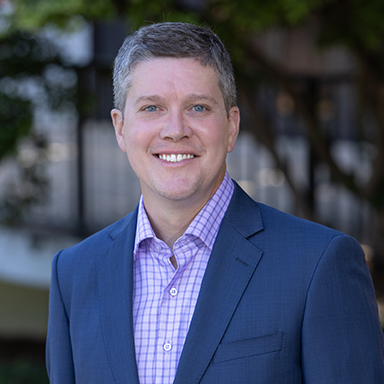 A professional headshot of a smiling man in a dark suit with a light purple checkered shirt, standing outdoors with blurred greenery in the background.