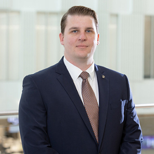 Professional headshot of a man in a dark suit and tie standing confidently in a well-lit interior setting.