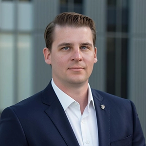 Professional headshot of a man in a dark suit and white shirt, standing against a modern architectural background.