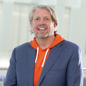 Smiling man in a blue blazer and orange hoodie, posing confidently in a modern indoor setting.
