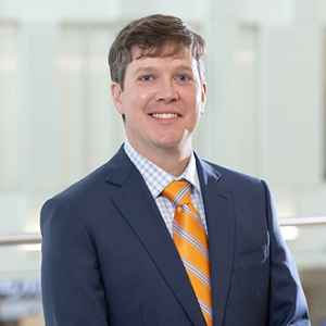 Professional headshot of a man in a suit with a blue jacket and orange tie, smiling for the camera.