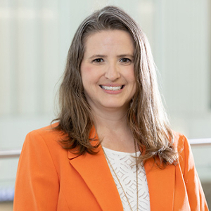 A professional portrait of a woman with long hair wearing an orange blazer and a white top, smiling confidently.