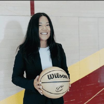 A woman in a blazer smiles while holding a Wilson basketball on a colorful gym floor.