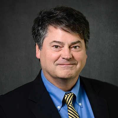 Professional headshot of a man in a suit and tie, smiling against a dark background.