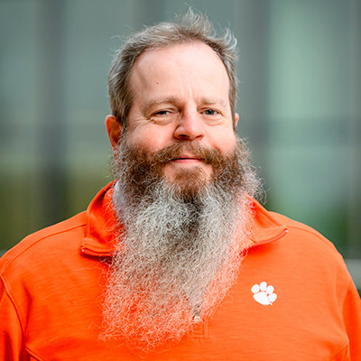 Close-up portrait of a smiling man with a long gray beard wearing an orange quarter-zip pullover with a small paw-print logo, outdoors with a blurred background.