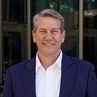 Smiling man in a navy blazer and white dress shirt standing in front of a modern glass building.
