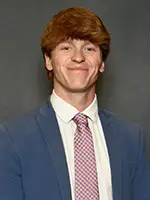 A young man in a blue suit and tie with a checkered pattern, smiling confidently against a neutral background.