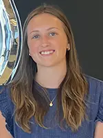 Smiling young woman with long brown hair, wearing a blue top, standing in front of a reflective surface.
