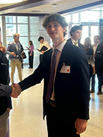 A young man in a suit shakes hands with another individual at a networking event, surrounded by attendees in a professional setting.