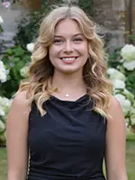 A smiling young woman with wavy blonde hair wearing a black dress, standing in front of a backdrop of white flowers.