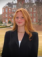 A young woman with long red hair wearing a black blazer, smiling confidently against a blurred campus background.