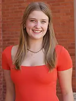 A young woman with long hair smiles while wearing a bright red dress, standing in front of a brick wall.