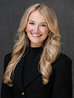 Studio headshot of a smiling woman with long blonde hair wearing a black blazer against a dark gray background.