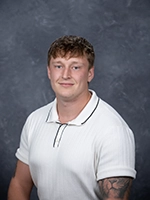 Professional headshot of a young man with short, curly hair, wearing a white polo shirt, posing against a gray background.