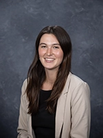 Professional headshot of a smiling woman with long brown hair, wearing a black top and light-colored blazer.