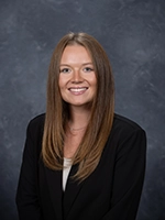Professional headshot of a woman with long hair, wearing a black blazer and smiling against a gray backdrop.