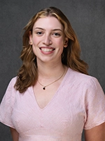 Studio headshot of a smiling woman with shoulder-length brown hair wearing a light pink top against a gray background.