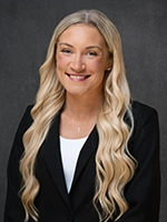 Professional headshot of a smiling woman with long wavy blonde hair wearing a black blazer over a white top, against a dark gray background.