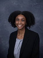 A professional headshot of a smiling woman with curly hair, wearing a black blazer and silver blouse.