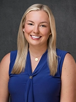 Studio headshot of a smiling woman with long blonde hair wearing a blue sleeveless blouse against a gray background.