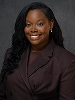 Professional headshot of a smiling woman with long dark hair, wearing a dark blazer against a gray background.