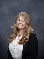 Professional headshot of a young woman with wavy blonde hair, wearing a white shirt and black blazer, smiling in front of a gray background.