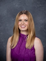 Professional headshot of a woman with long blonde hair, wearing a purple blouse, set against a gray background.