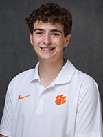 Smiling young man in a white polo shirt with an orange paw logo, posed against a dark gray studio backdrop.