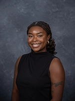Headshot of a young woman with braided hair, wearing a sleeveless black top, smiling against a dark gray background.