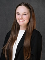 Studio headshot of a smiling woman with long brown hair wearing a black blazer over a white top against a gray background.