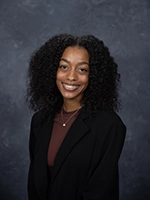 A professional headshot of a smiling young woman with curly hair, wearing a black blazer over a brown top.