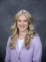 Professional headshot of a woman with long, wavy blonde hair, wearing a light purple blazer and a white top, smiling against a gray background.
