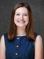 Smiling woman with shoulder-length brown hair wearing a sleeveless dark blue top, posed against a gray studio background.