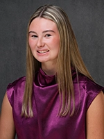 Studio portrait of a smiling young woman with long blonde hair wearing a purple sleeveless top against a gray backdrop.