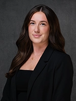 Studio headshot of a woman with long dark hair wearing a black blazer, smiling against a gray background.