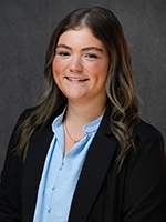 Studio headshot of a smiling woman wearing a black blazer over a light blue collared shirt against a dark gray background.