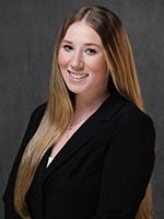 Professional studio headshot of a smiling woman in a black blazer against a dark gray background.