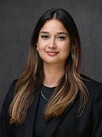 Professional headshot of a woman with long brown hair, wearing a black blazer, against a gray studio background.