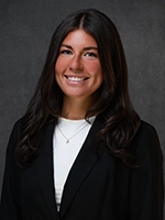 Studio portrait of a smiling woman with long dark hair, wearing a black blazer over a white top against a dark gray background.