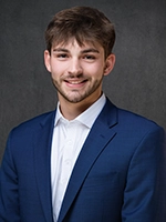 Studio headshot of a smiling man wearing a blue suit jacket and white shirt against a dark gray background.