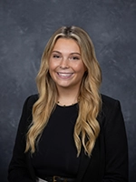 Professional headshot of a smiling young woman with long blonde hair, wearing a black blazer over a black top.